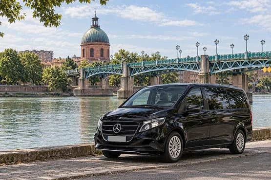 Van Mercedes noir au bord d’un fleuve, devant un pont et un dôme en briques.