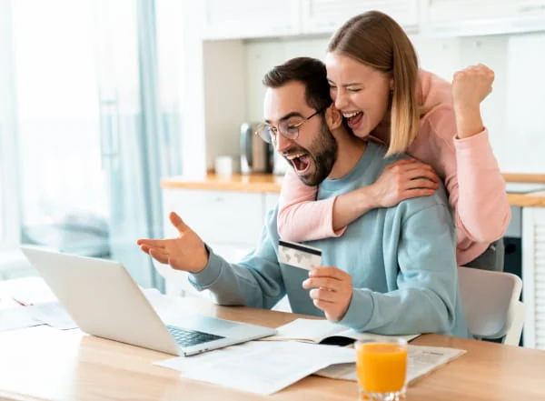 Homme tenant une carte bancaire devant un ordinateur portable, avec une femme souriante derrière lui.